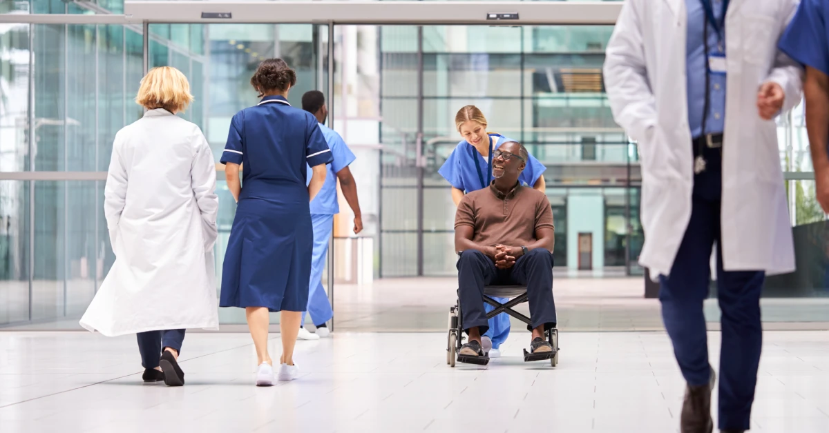 Female-Nurse-Wearing-Scrubs-Wheeling-Patient-In-Wheelchair